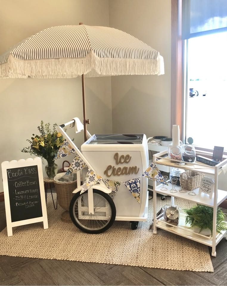 The Belle ice cream cart set up indoors for a reception, with a striped fringed umbrella, florals and a chalkboard flavor menu